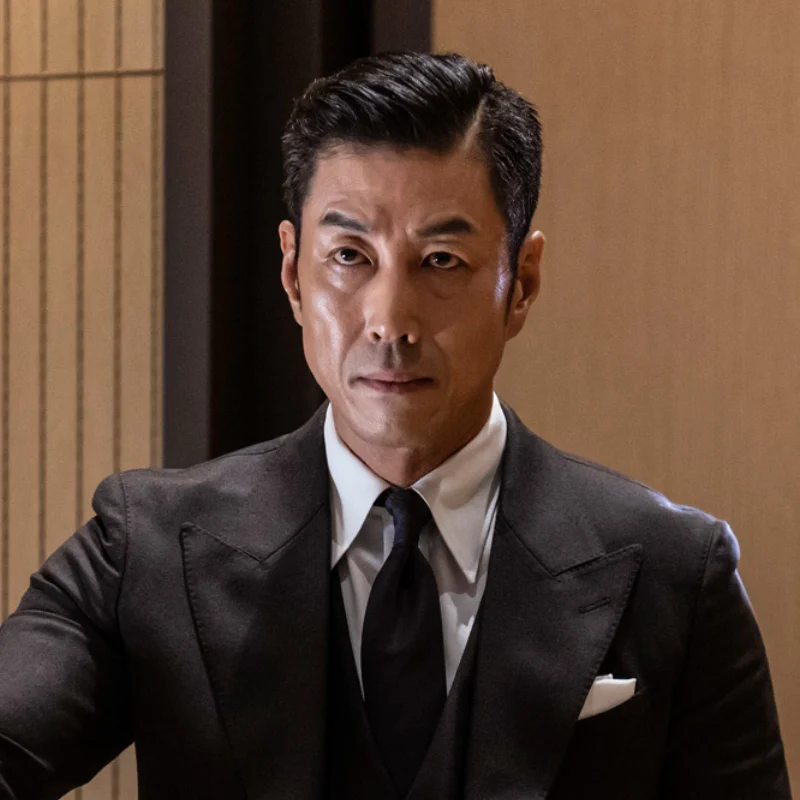 어느 오피스 실내를 배경으로 머리를 단정히 올리고 검은 슈트를 입고 서 있는 드라마 7인의 탈출의 강기탁 캐릭터