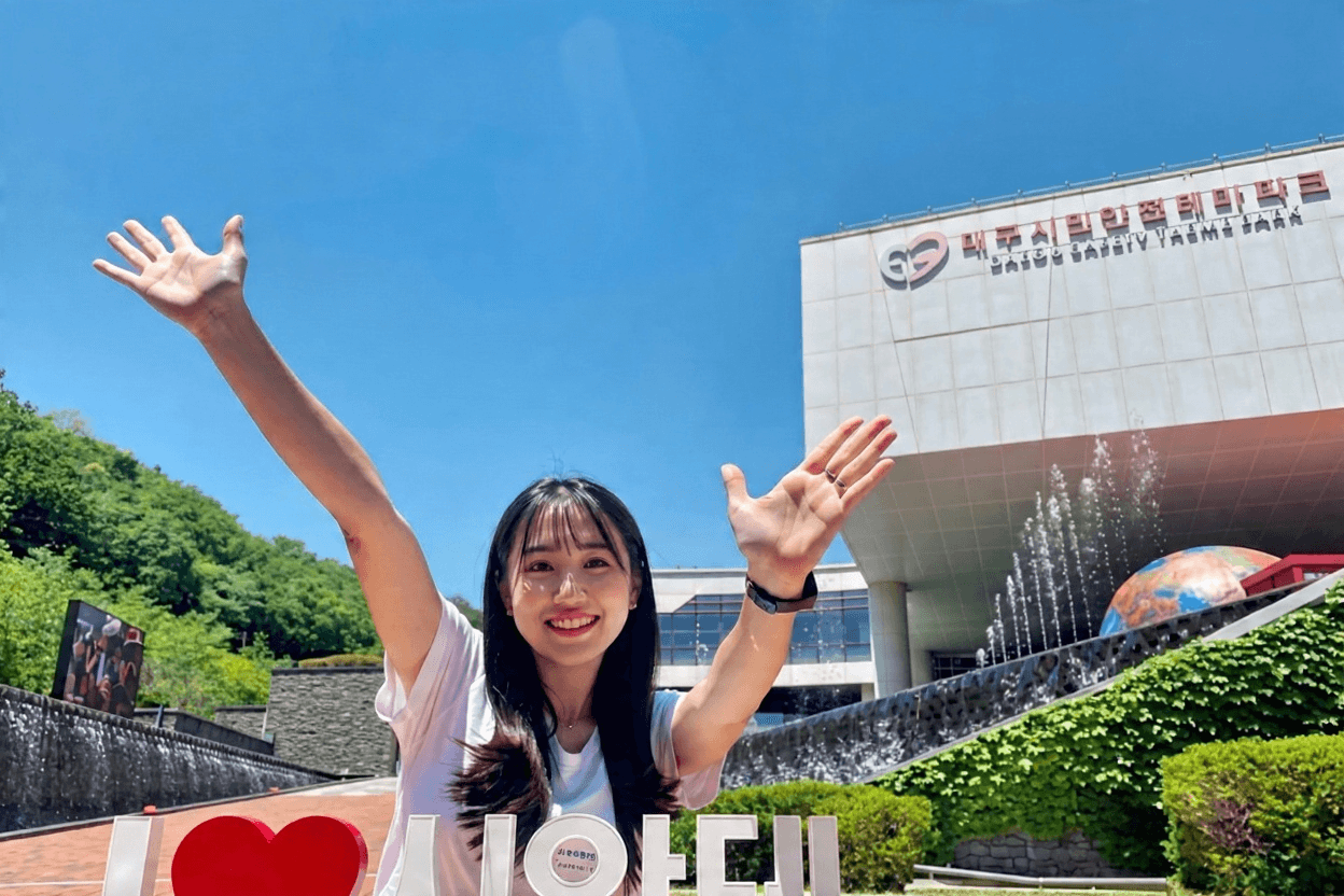 대구 시민안전테마파크 건물 외관 앞 I LOVE 포토존에서 기념사진을 찍는 여성, 파란 하늘 아래 분수대 물줄기와 지구본 조형물, 초록빛 조경과 DAEGU SAFETY THEME PARK 간판이 보이는 주간 전경