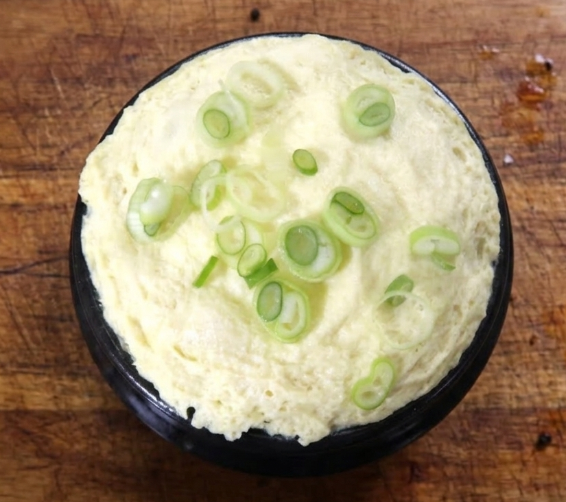 사용감 있는 빈티지 나무 도마 위에 놓인 검은색 뚝배기 폭탄 계란찜과 송송 썬 대파 고명 사진.