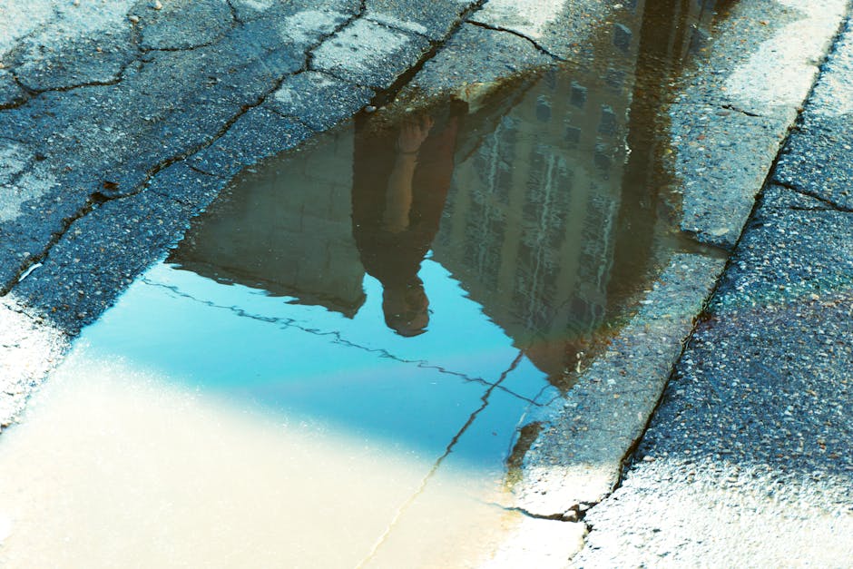 Reflection of a person and buildings in a puddle on cracked asphalt, capturing urban textures.