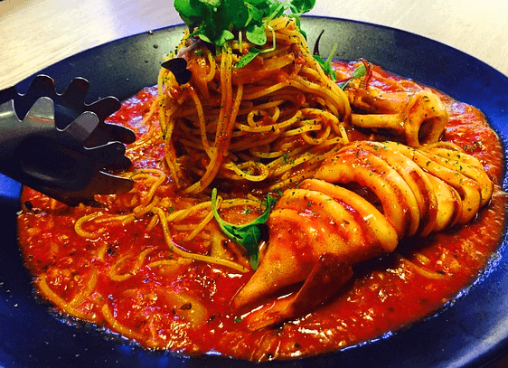 동그란 검정 접시에 붉은 색의 토마토 스파게티가 있도 오른 쪽에는 동그랗게 썰어진 통오징어 한마리가 있다.