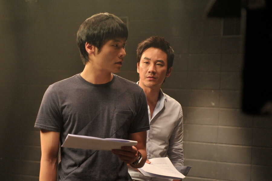 대본을 연기하는 남자와 그 뒤에서 그를 바라보는 대본 든 남자