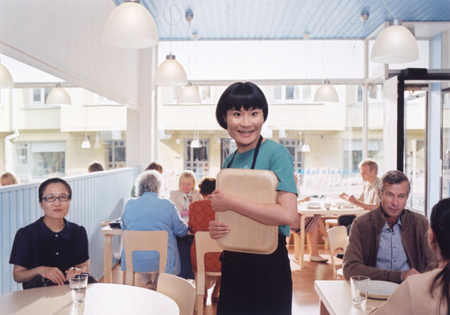손님으로 테이블이 꽉 찬 식당을 배경으로 여자 주인공이 웃고 있는 모습