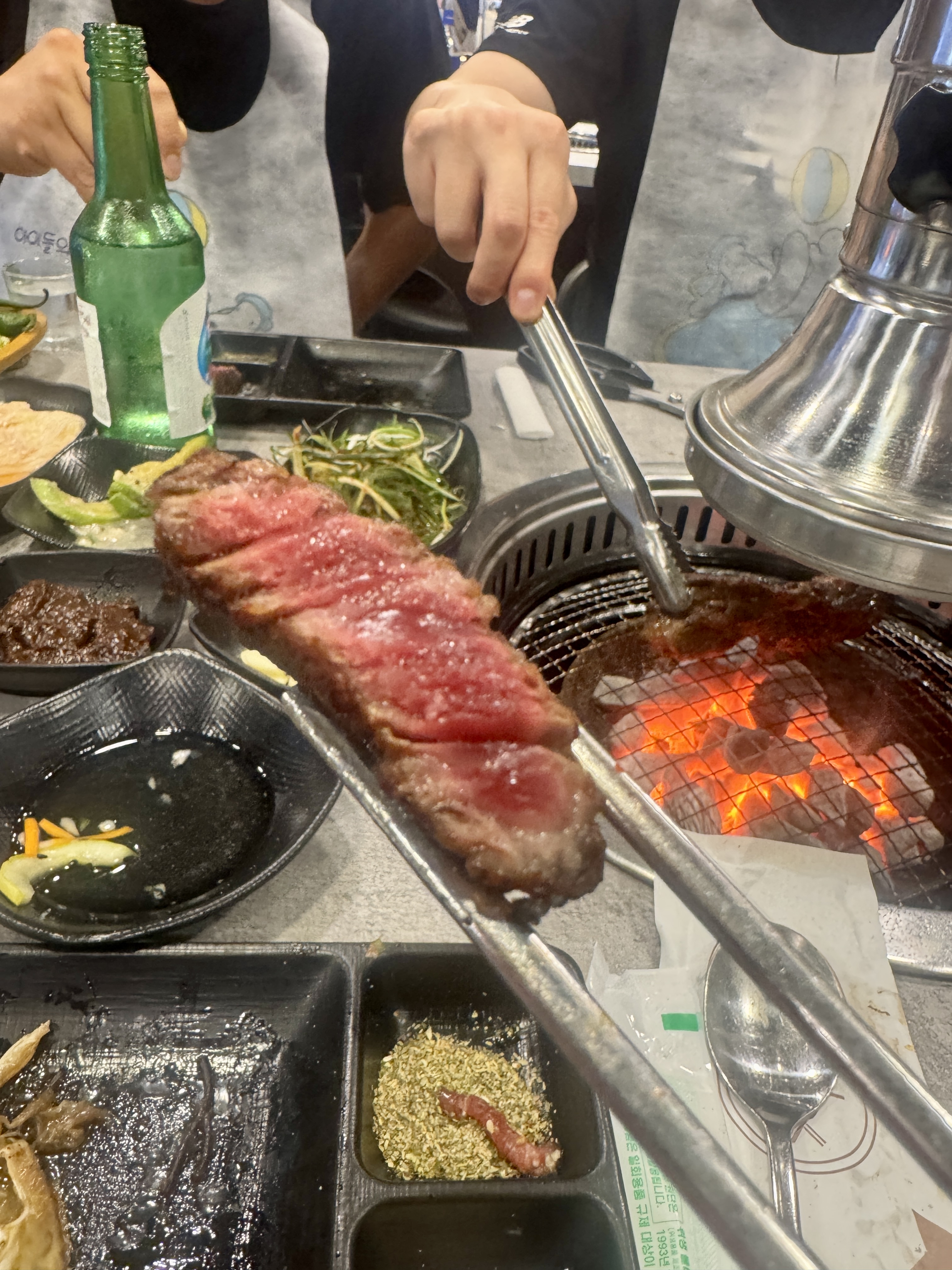 삼거리 명품한우에서 마무리로 먹는 듯한 구운 한우 한 점. 마블링이 살아 있고 육즙이 흐르며 배경엔 숯불과 소금&amp;#44; 양념장이 보인다.