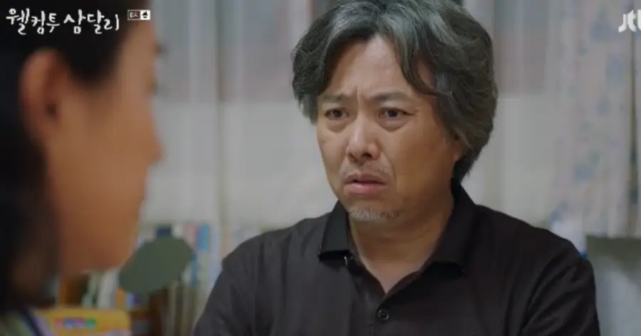 웰컴투삼달리- 검정색 셔츠에 히끗히끗한 머리로 걱정스러운 얼굴로 상대방과 대화하는 모습