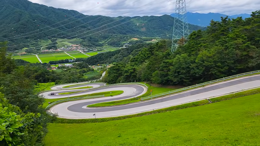 함양 지안재 S자 고갯길 전경, 초록 초지와 굽이진 도로, 여름 구름 하늘
