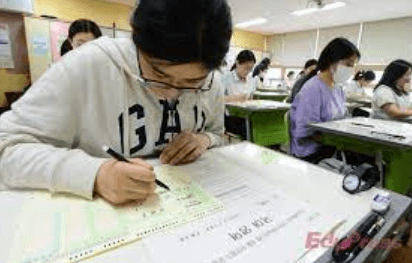입시제도- 흰색시험지 위 검은색 펜을 들고 흰색 후드티를 입고 안경을 쓴채 시험을 치르는 여학생