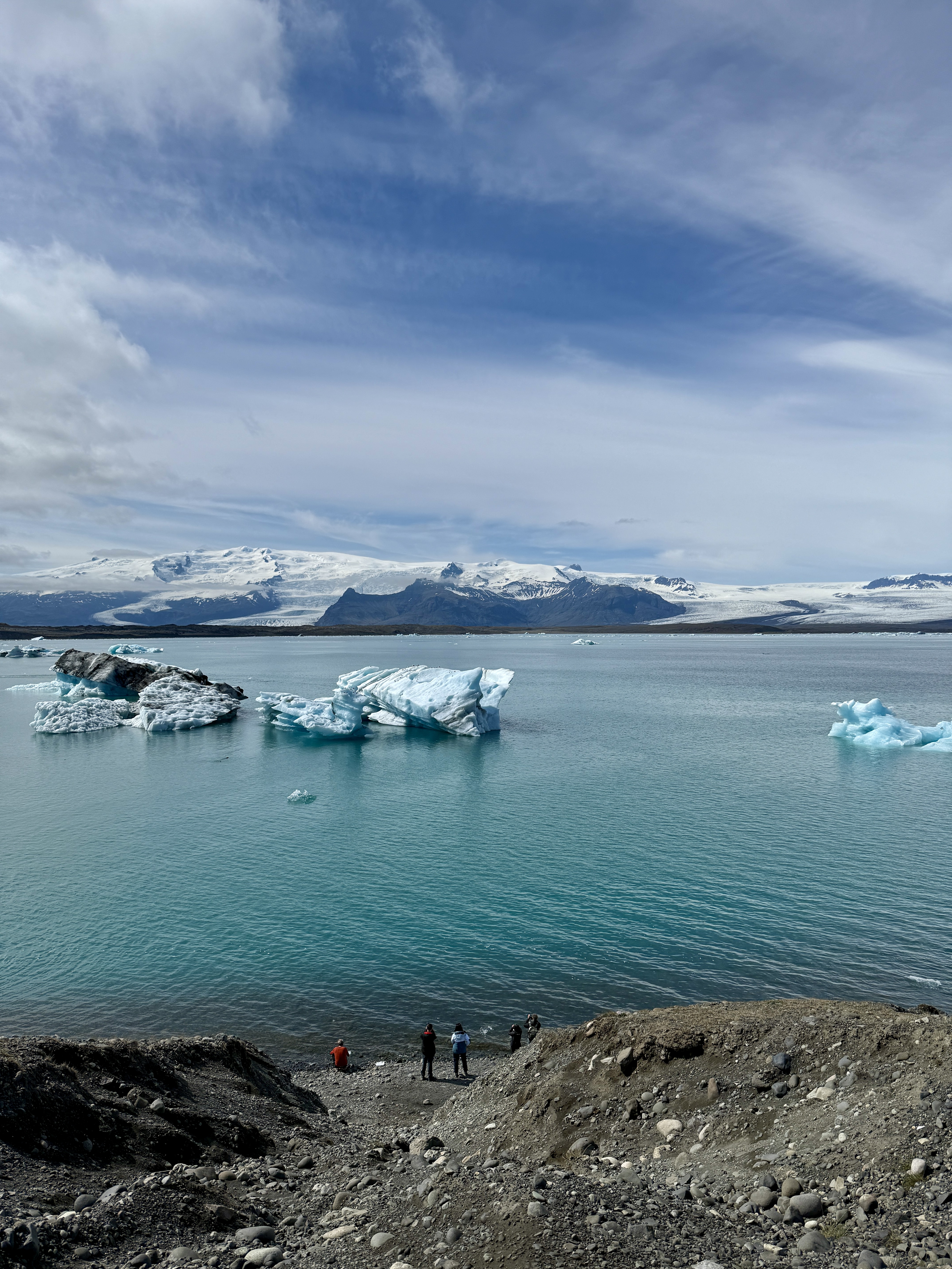 요쿨살론 빙하 호수의 장관. 청록색 물 위에 크고 작은 빙하 조각들이 떠 있고, 멀리 설산과 빙하가 펼쳐져 있음. 호숫가 아래쪽에는 빙하의 웅장함을 느끼게 하는 작은 규모의 관광객들이 서 있음.