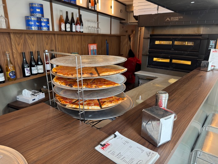 An indoor view of MIKE pizza bar showing a layered pizza cooling rack and a large Moretti Forni oven