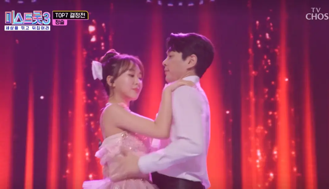 미스트롯3- 빨간무대 앞 분홍색 드레스를 입은 정슬이 흰색 와이셔츠를 입은 남자의 어깨에 손을 얹고 함께 춤추는 장면