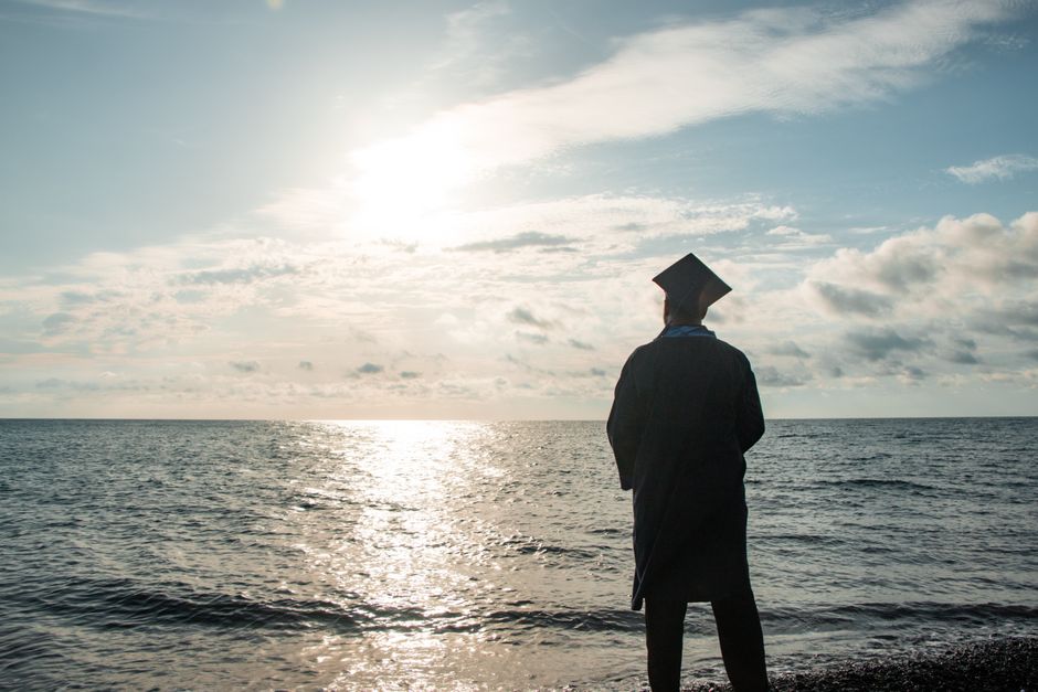 Graduate in cap and gown contemplating the future by the sea at sunset.