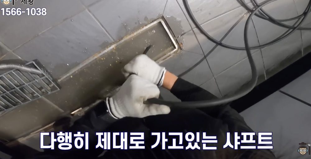 작업자가 바닥 트렌치 배수구 안으로 샤프트 장비를 넣어 깊은 배관 막힘 구간을 청소하는 장면