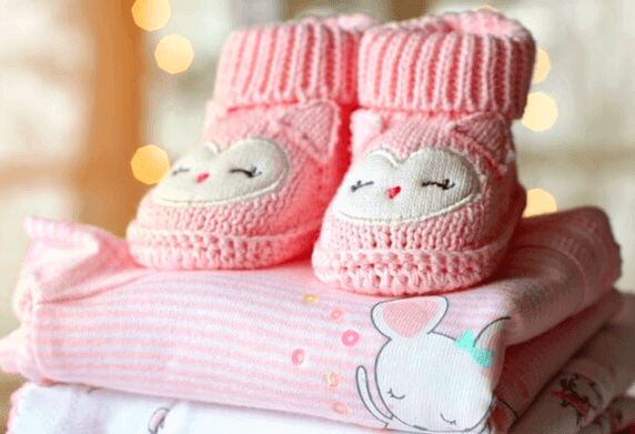 분홍색 아기 옷이 잘 개어져 있고 옷 위에는 분홍색 아기 털신발이 있는데 신발 표면에는 웃는 아기 미소가 그려져 있다.