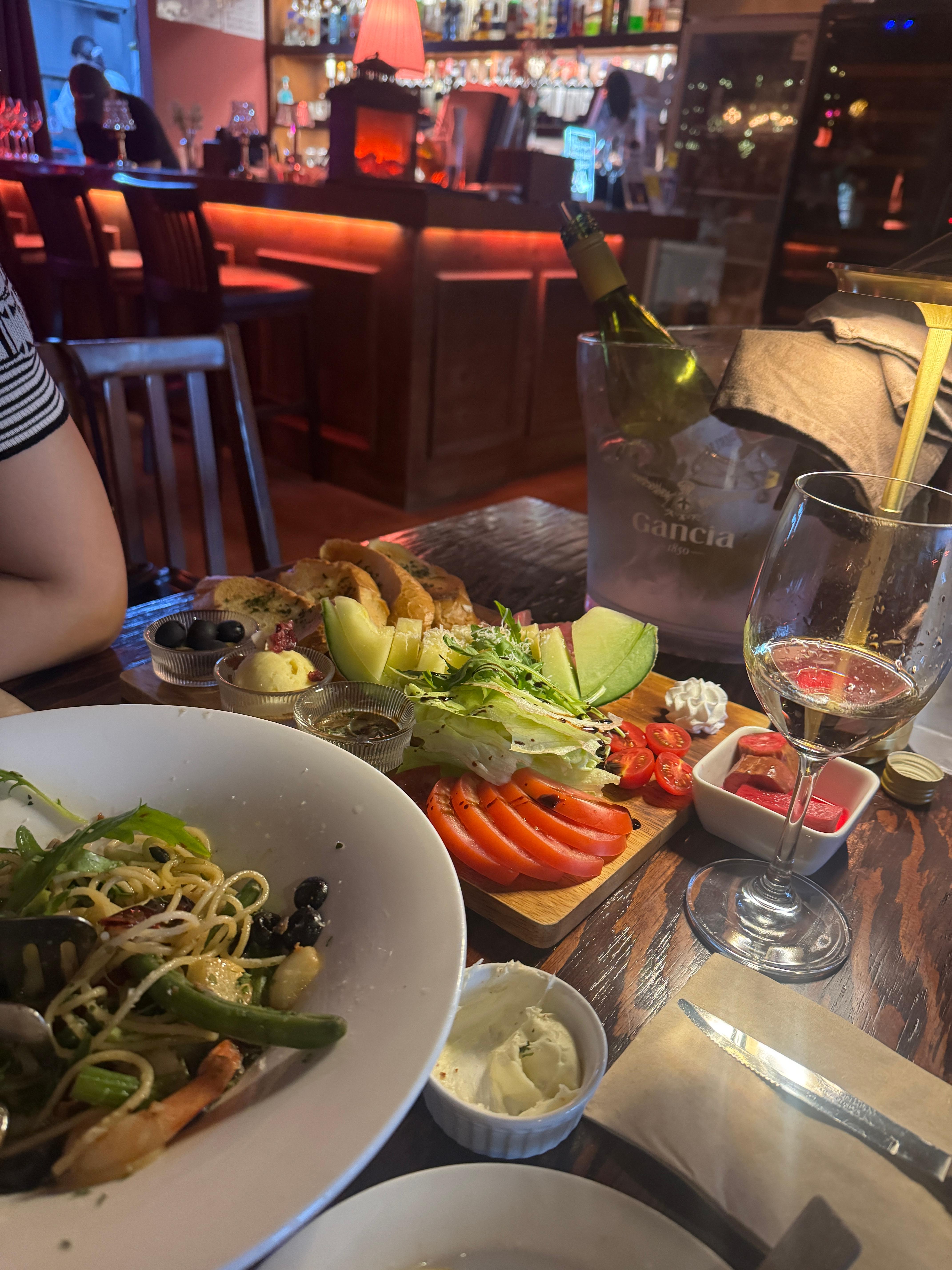 논현동 와인바에서 주문한 화이트 와인, 하몽 플레이트, 쉬림프 알리오 올리오 파스타가 테이블 위에 놓여 있다. 토마토와 치즈, 채소가 풍성하게 담긴 안주와 와인잔이 아늑한 조명 아래 어우러져 있다.