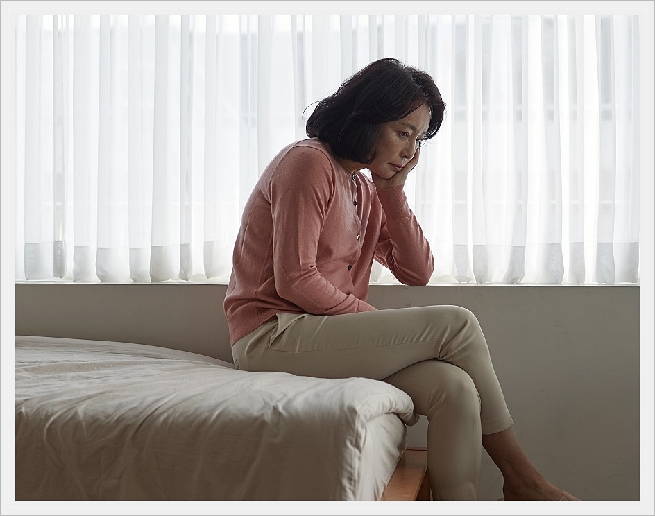 침대에 걸터 앉아 다소 가라앉은 표정을 짓고 있는 중년 여성의 모습
