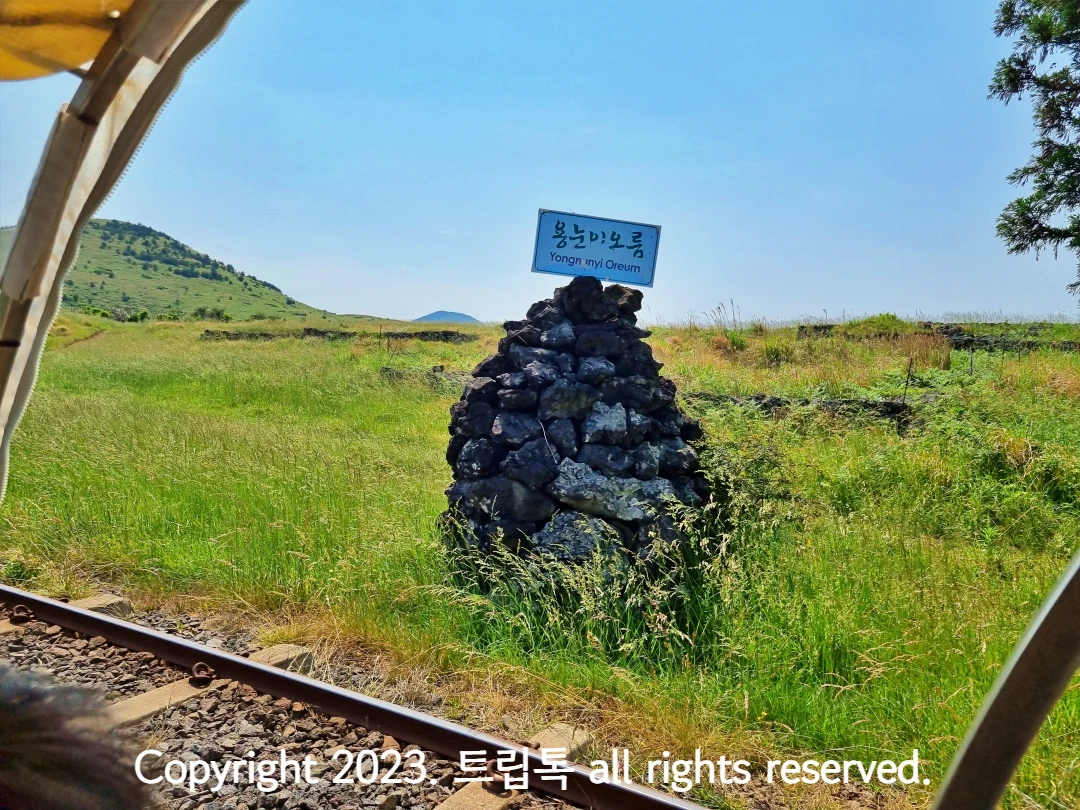 현무암 돌탑에 용눈이오름이라고 적혀있고 그 왼쪽에 용눈이 오름이 있다.