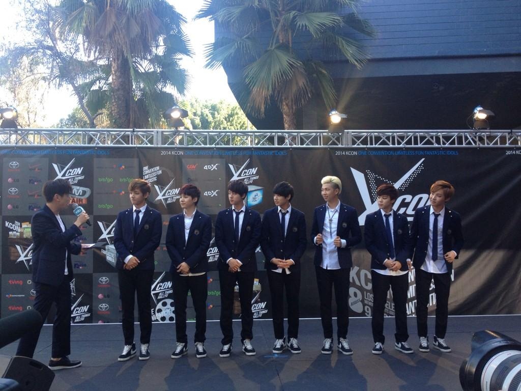 BTS and host Eric Nam standing in dark suits on the KCON 2014 red carpet, with event banners and palm trees behind them.