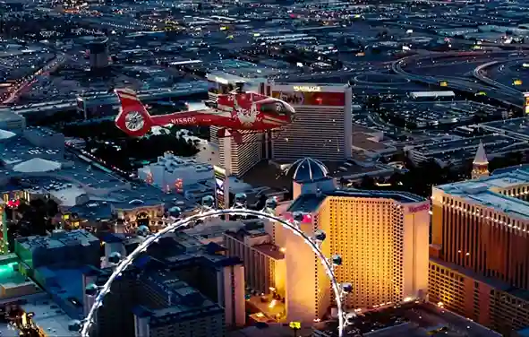 라스베이거스 스트립의 화려한 야경 위로 빨간색 헬기가 비행하는 모습. 호텔 건물과 관람차가 보인다.