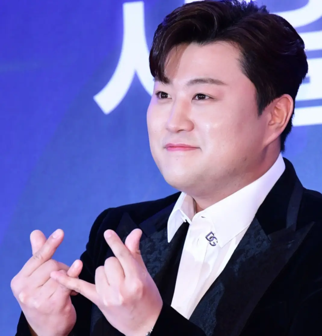 파란배경 검은옷을 입은 김호중이 미소지으며 손하트를 그리는 모습