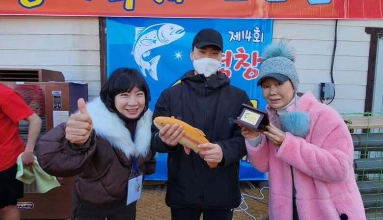 검정색 옷과 모자를 쓴 한 남성이 가운데 서있고 양쪽에는 여성이 각각 서있는 모습으로 왼쪽 여성은 오른손으로 엄지를 치켜세우고 웃고 있고 오른쪽 여성은 황금송어를 잡은 트로피를 내보이고 있고 가운데 남성은 황금색 송어 인형을 들고 있는 모습