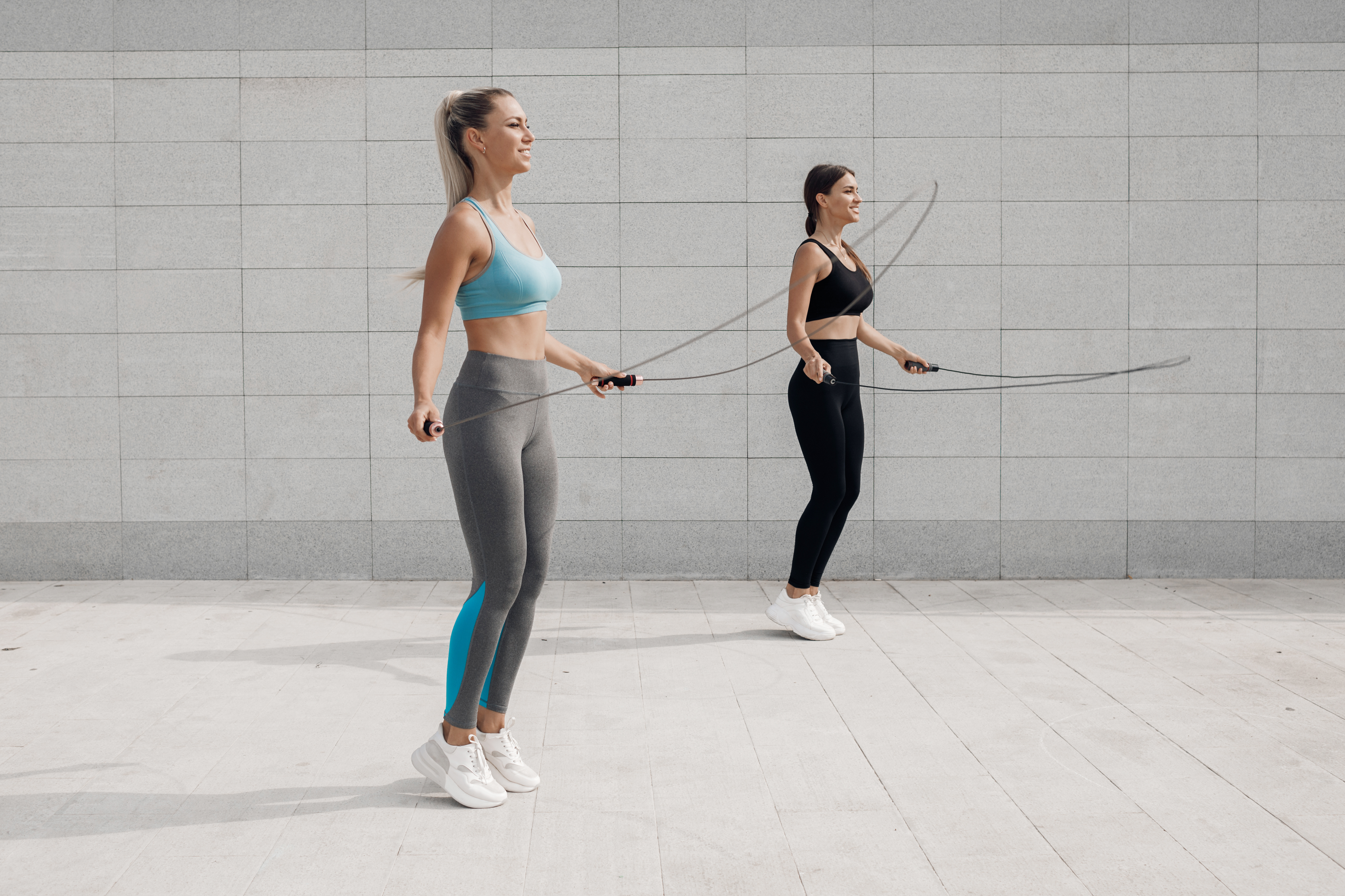 2명의 여성이 운동복을 입고, 실외의 길 위에서 나란히 서서 줄넘기를 하고 있는 사진(two-friends-fitness-outdoor)