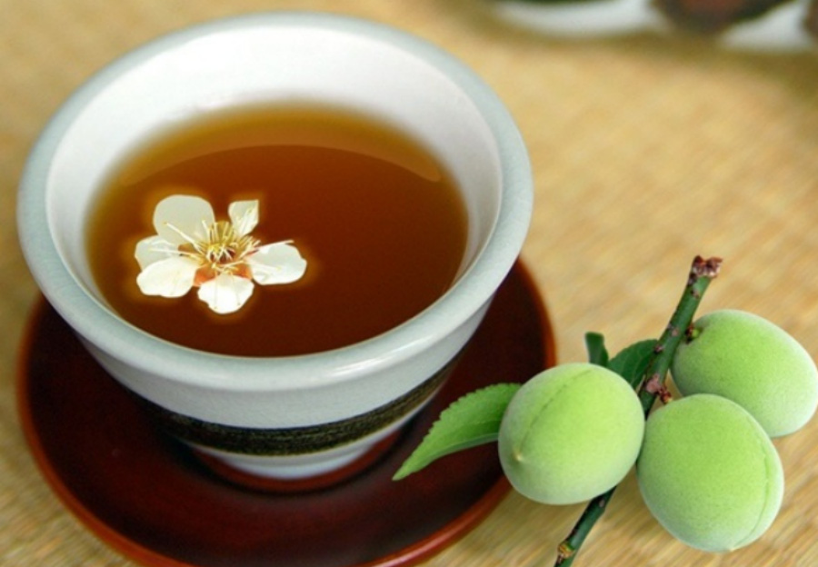 대나무 바닥에 매실이 3개가 달려 있는 줄기와 매실을 끓여 만든 매실차에 매실 꽃을 1개 담은 찻잔세트를 같이 놓아두고 찍은 사진