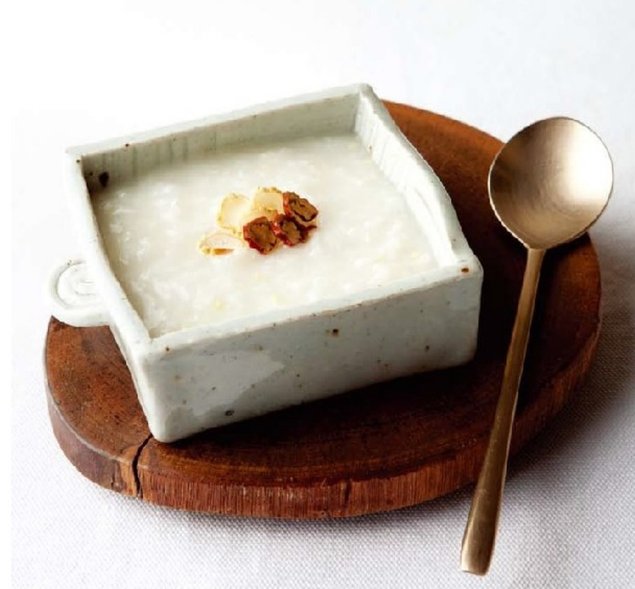 쌀과 인삼을 넣고 끓여서 만든 인삼죽을 네모난 그릇에 가득 담아 놓고 대추를 썰어서 놓은 것을 하얀 바닥 위에 동그란 나무 받침대 위에 쇠숟가락과 함께 놓아두고 찍은 사진