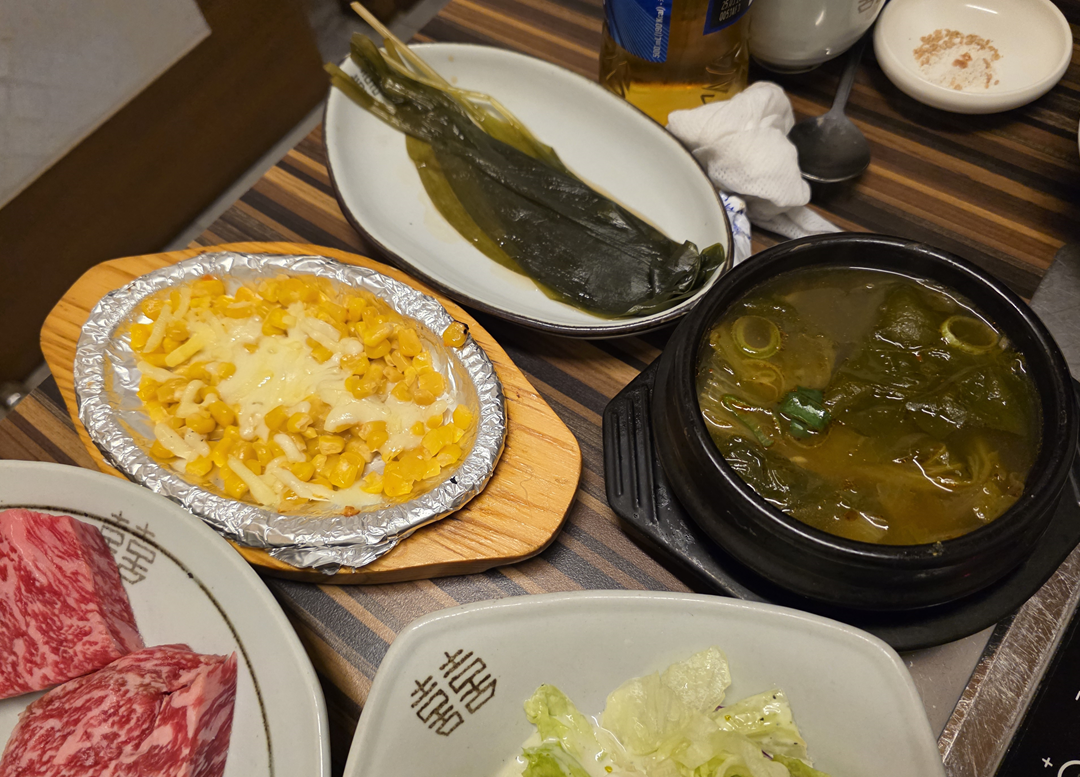 치즈를 올린 콘버터와 명이 나물&amp;#44; 미역국이 함께 놓인 한우 식사 반찬 구성 전경