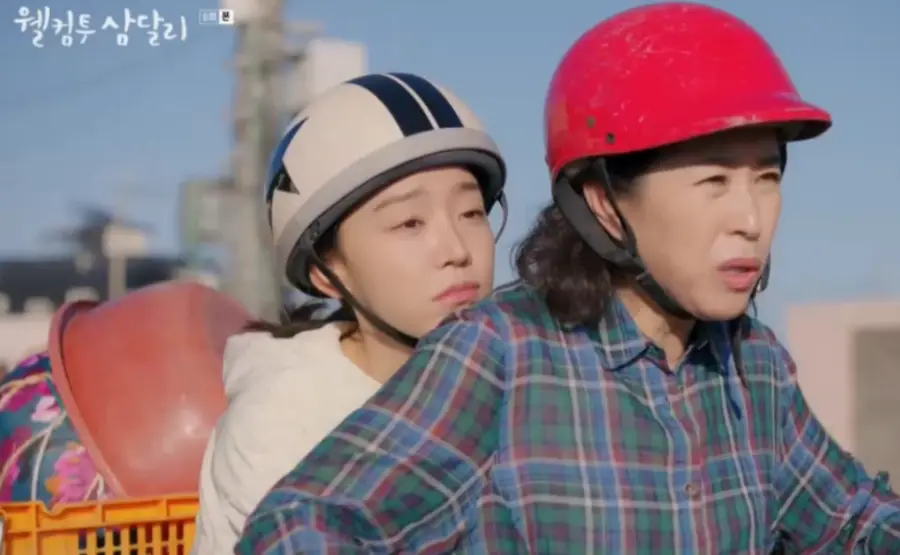 웰컴투삼달리- 빨간헬맷을 쓰고 파란색 연두색 격자무늬 셔츠를 입고 뒤에 흰헬맷을 쓴 삼달이를 태우고 가는 고미자