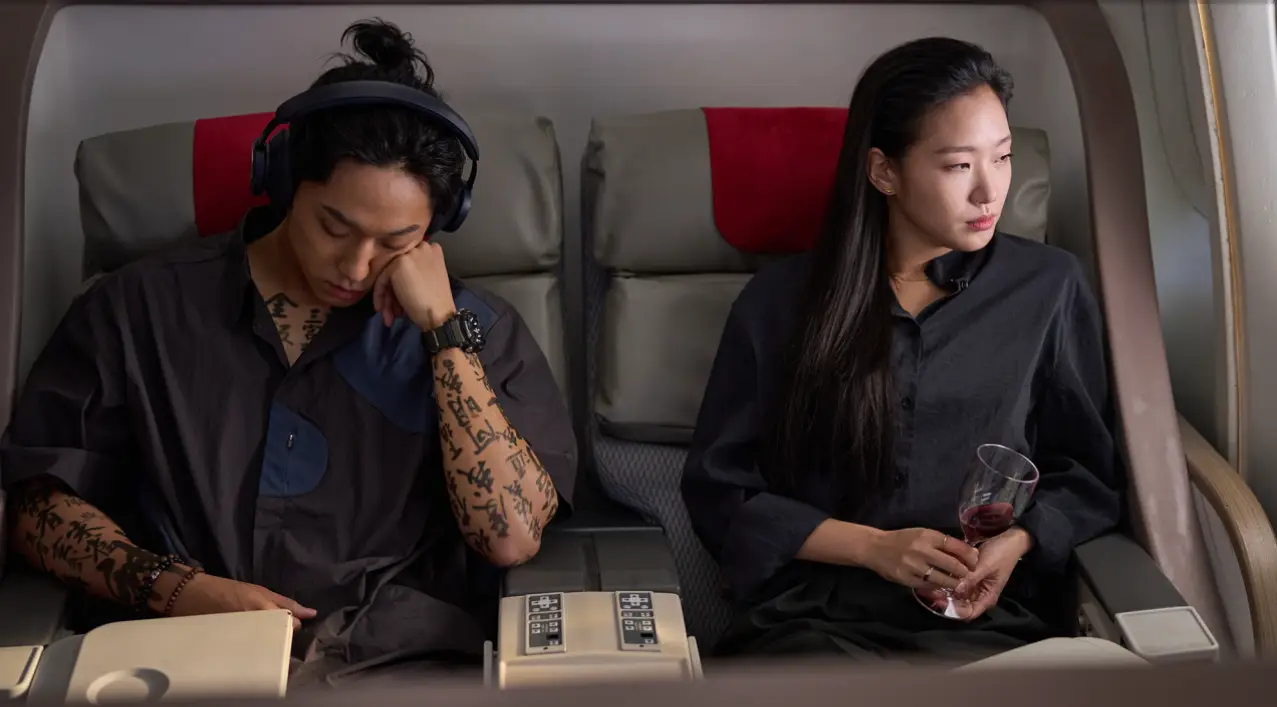 파묘- 비행기 안 회색의자에 안자 헤드폰을 쓰고 왼손으로 머리를 괸채 잠을자는 이도현과 검은 블라우스에 와인잔을 들고 밖을 바라보는 김고은