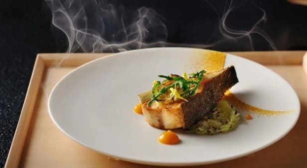 흰 색 접시에서 연기가 피어오르고 접시위에는 생선구이가 놓여져 있다.