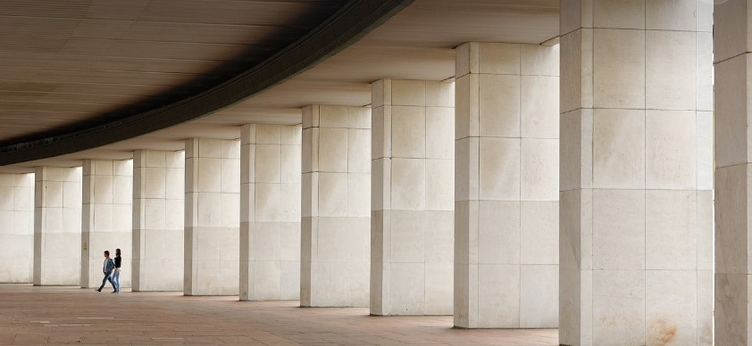 많은 기둥이 있는 건축물 사이로 두사람이 걸어가고 있는 사진