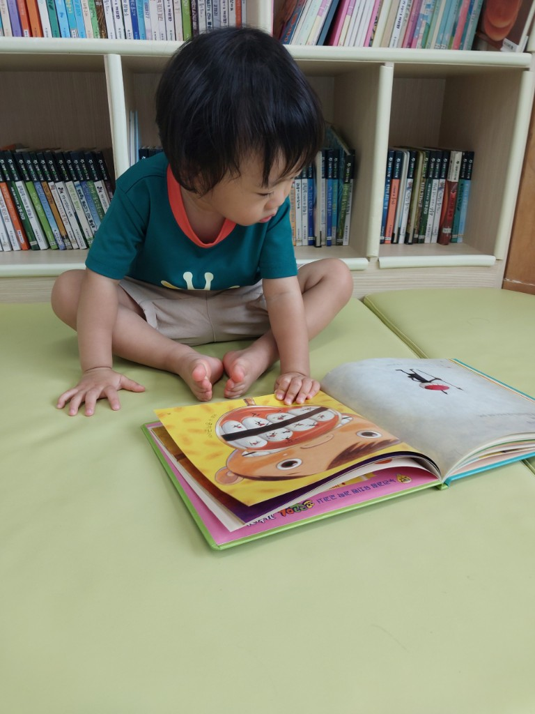 집 책장 앞 바닥에 앉아 치아 그림책을 진지하게 탐색하며 읽고 있는 30개월 아이 모습