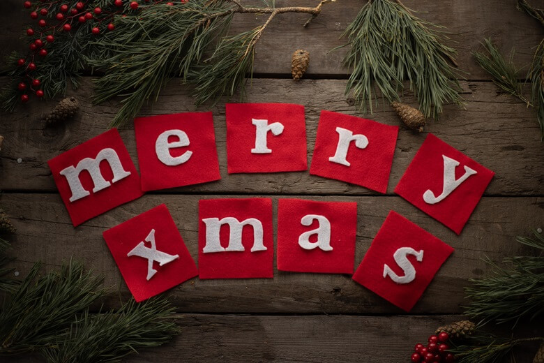 나무로 된 바닥위에 사각형 모양의 빨간색 천위에 하얀색 글자를 각각 한글자씩 모아서 펼쳐둔 영어로된 메리 크리스마스 글자와 그 주변에 소나무 가지와 솔방울과 빨간열매로 장식된 사진