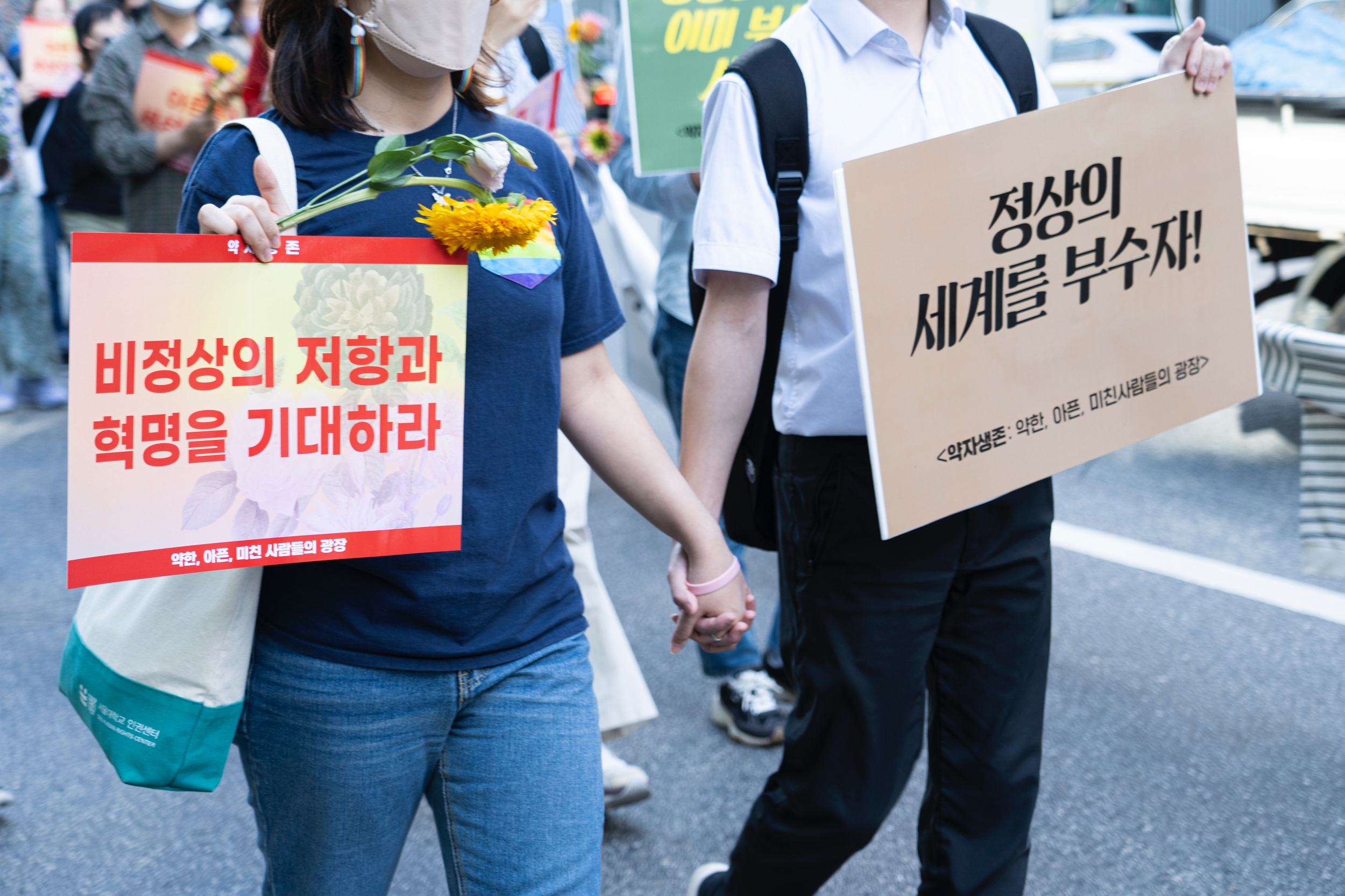 '비정상의 저항과 혁명을 기대하라'와 '정상의 세계를 부수자' 피켓을 든 시민들이 서로 손을 잡고 행진하고 있다.