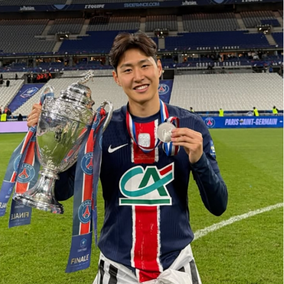 축구 유니폼을 입은 선수가 경기장에서 트로피와 은메달을 들고 미소 짓고 있다. 유니폼에는 파리 생제르맹(PSG) 로고가 보이며, 배경은 관중석이 비어 있는 스타디움이다.