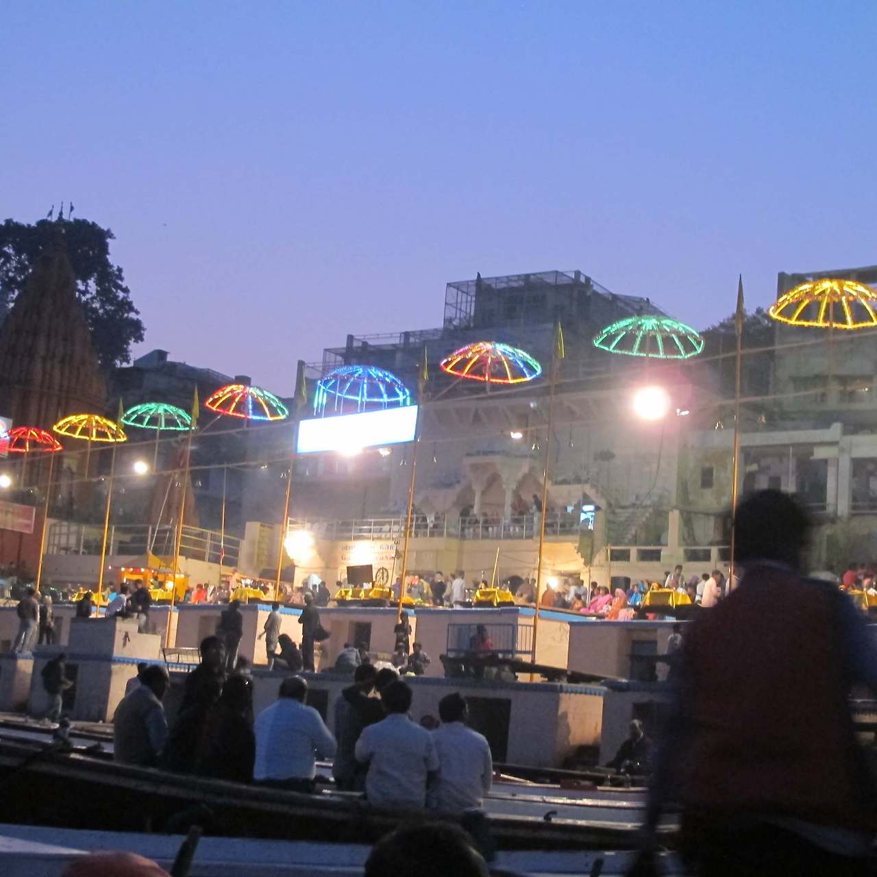 가트의 계단 위로 화려한 네온 조명이 장식된 파라솔들이 줄지어 있고 사람들이 앉아 있는 밤 풍경