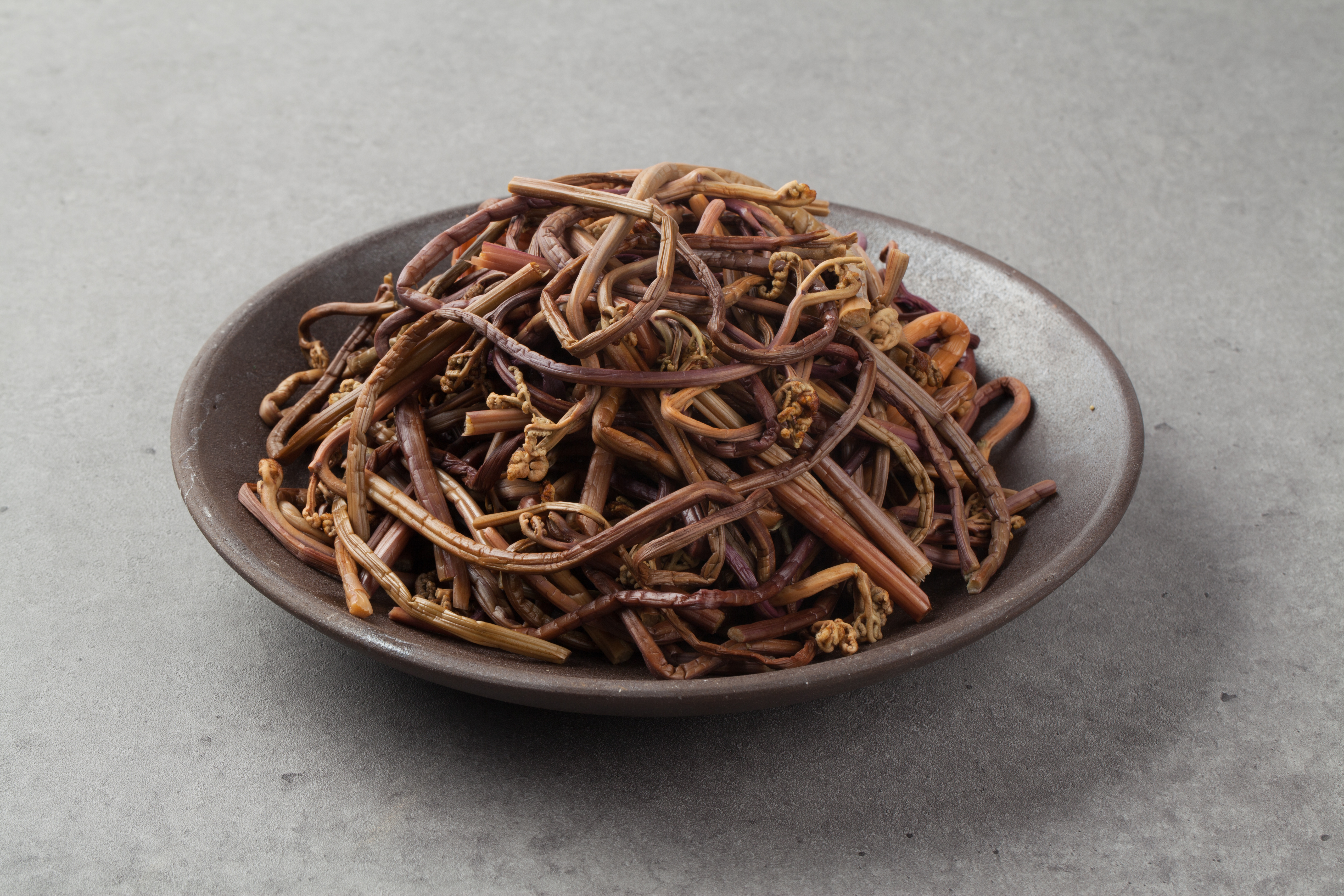 야생에서 채취를 한 고사리를 잘 세척하고 삶아서 회색의 바닥 위에 동그랗고 고동색의 유리 그릇 위에 수북하게 담아 놓고 찍은 사진