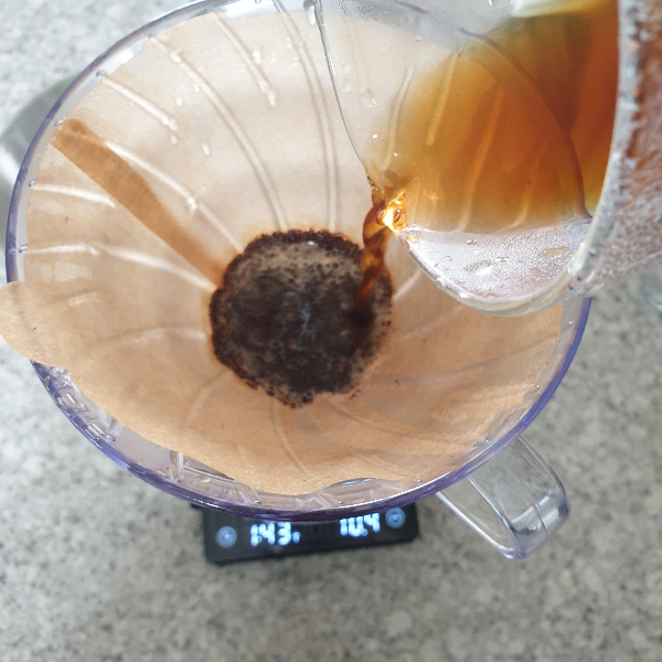 저울 위에 드리퍼가 올려져 있다. 드리퍼 안 쪽에는 젖은 커피 가루가 있고 서버로 추출된 커피를 드리퍼에 붓는 중