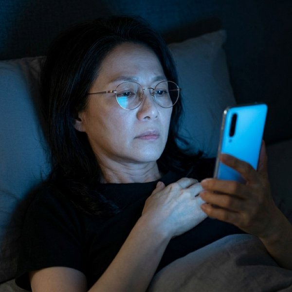 어두운 밤 침대에 누워 스마트폰 블루라이트를 얼굴에 쬐며 잠들지 못해 피곤해하는 50대 한국 여성의 1:1 비율 실사 사진.