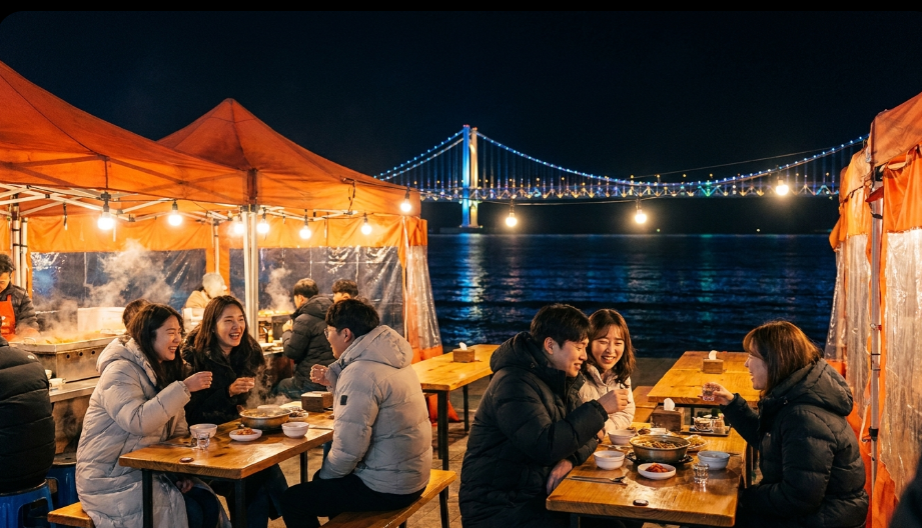 밤바다 야경 아래 조명이 켜진 야외 포차에서 즐겁게 대화하는 사람들