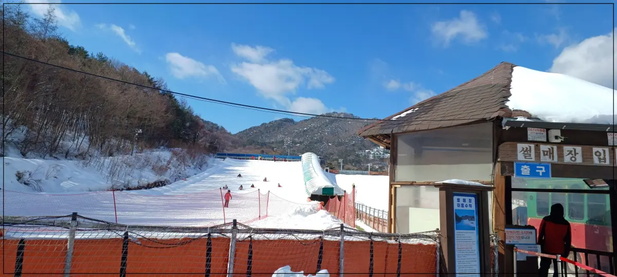 눈썰매장에서-아이들이-내려오고-있음-멀리-하늘과-산이-보임-앞에는-썰매장입구-출구라고-써있고-안전요원이-서있음