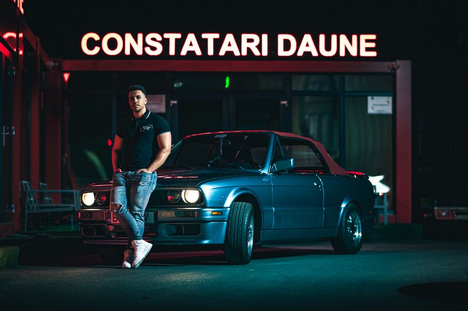 A fashionable man leans on a vintage blue car under neon lights at night.