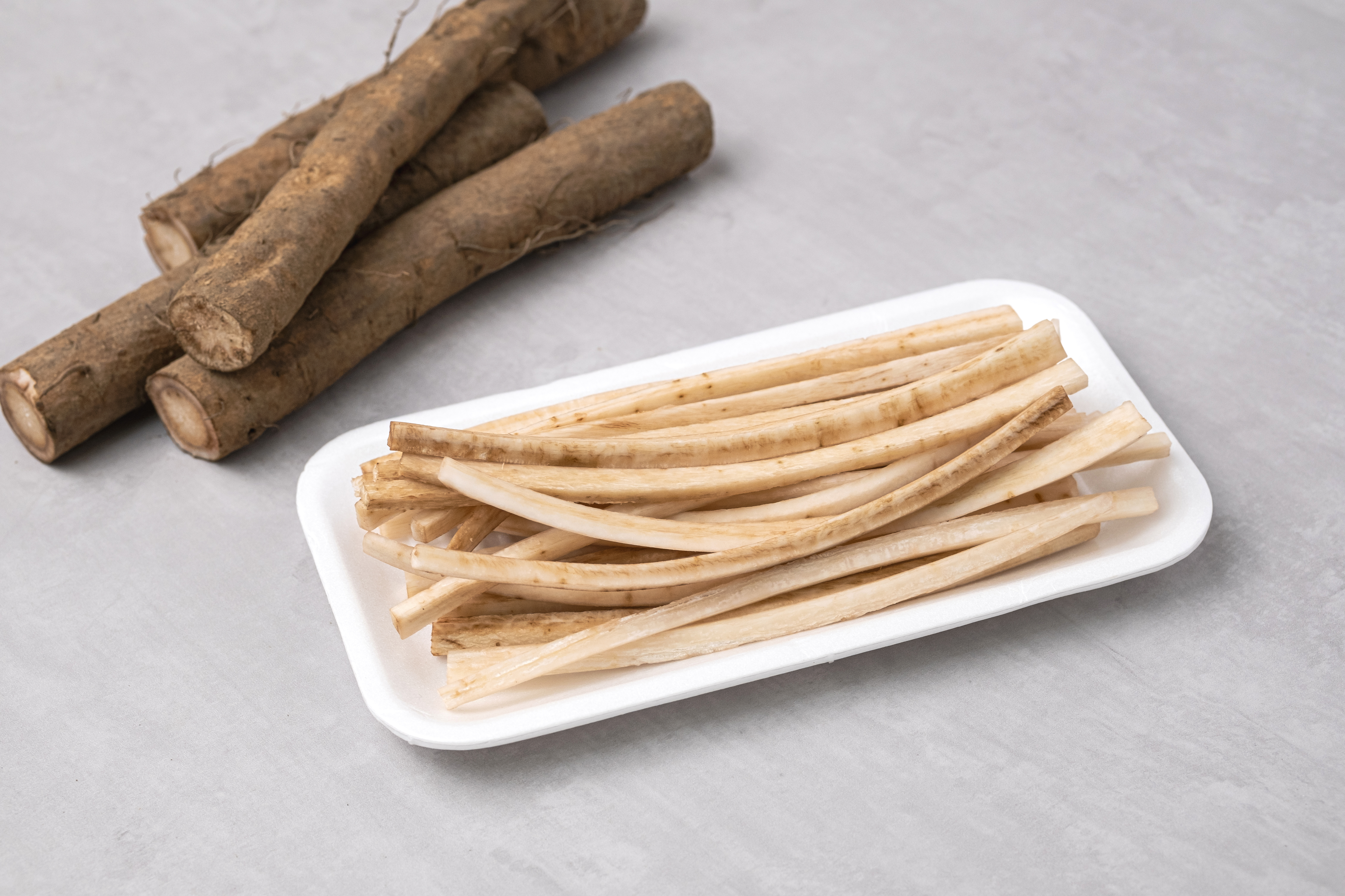 땅에서 캐낸 우엉 뿌리를 토막내서 바닥에 두고, 그 밑에 플라스틱 쟁반 위에 우엉의 껍질을 까내고 세로로 길게 잘라서 놓아 둔 사진