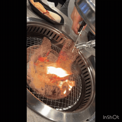 삼거리 명품한우에서 두툼한 소고기를 숯불 위에서 굽고, 가위로 자르는 장면. 불꽃이 올라오고 석쇠 위에서 고기가 익고 있다.