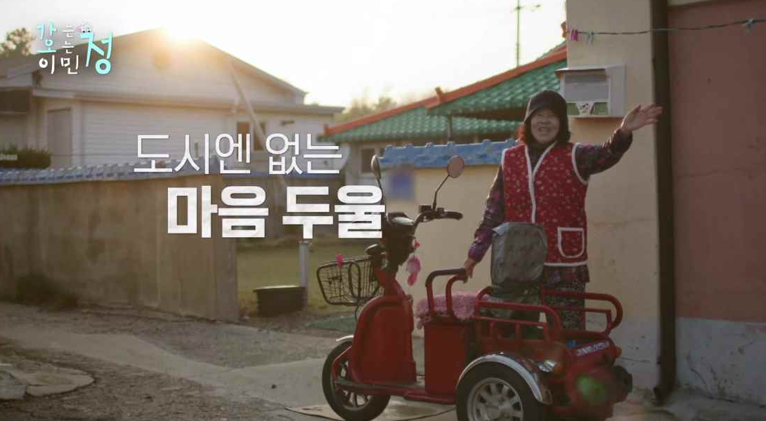 동네 마을 주민 집에 빨간 오토바이 옆에서 아주머니가 웃으면서 손 흔드는 장면에 도시엔 없는 마음 두울 자막이 있는 스틸컷