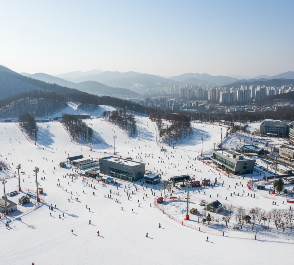 맑은 겨울날, 드론으로 촬영한 수도권 스키장 전경. 설산과 도시 풍경을 배경으로 슬로프에서 스키와 스노보드를 즐기는 사람들과 스키장 시설이 보인다.