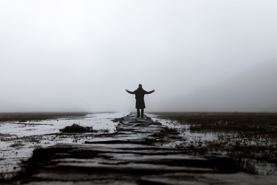 A solitary figure stands on a misty stone pathway in a rural area surrounded by fog.