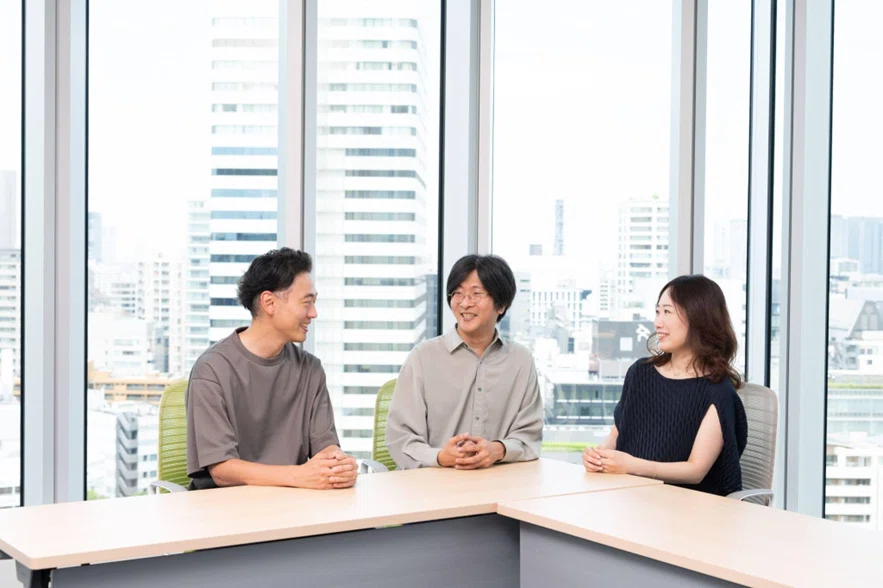 스즈키, 오하시, 마스다가 담소를 나누는 사진. 오피스의 창문을 배경으로 촬영하고 있다.