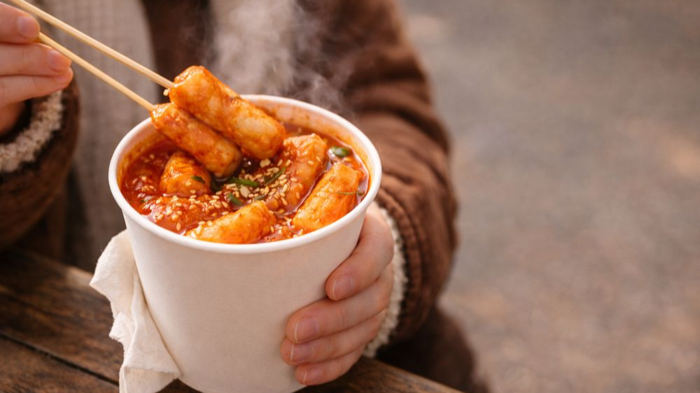 손으로 종이컵을 들고 떡볶이를 젓가락으로 집어 먹는 따뜻한 겨울 길거리 간식 장면